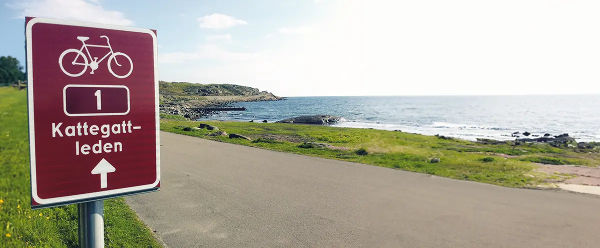 Brun skylt med texten "Kattegattleden" står bredvid en cykelväg vid havet.