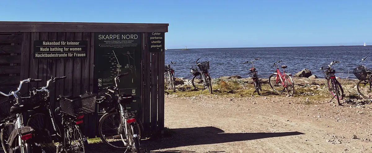 Trävägg med skylt där det står nakenbad för damer och Skarpe nord. Intill står cyklar uppställda och havet är i bakgrunden.