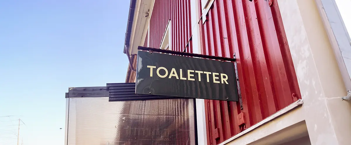 Skylt med texten "Toaletter" som sitter på en röd fasad.