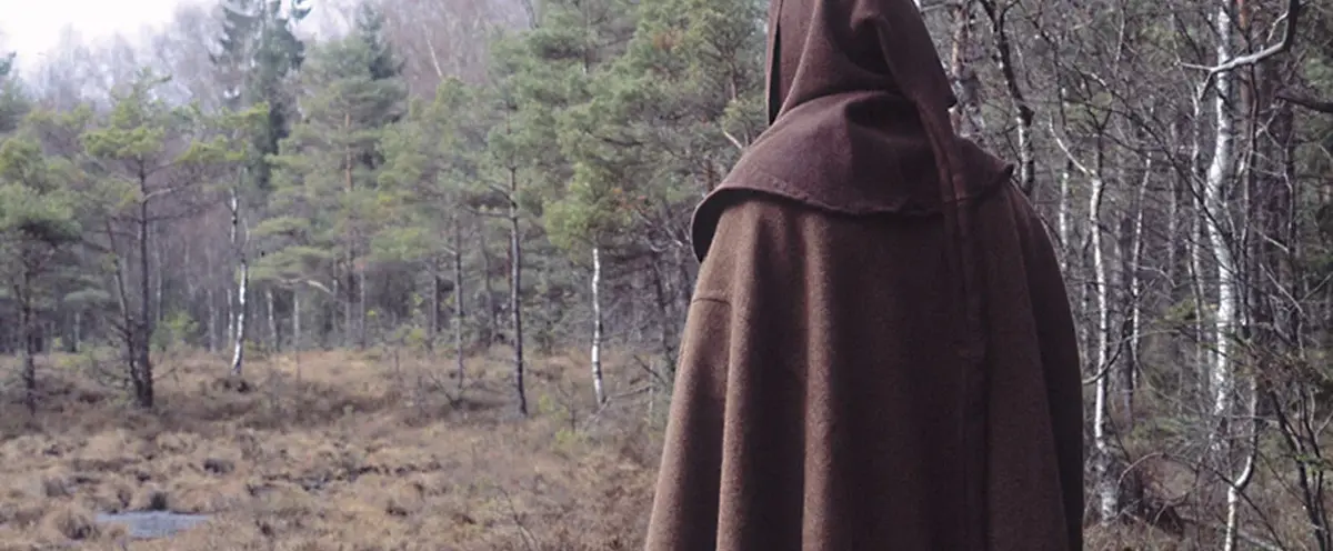 En person med brun klädnad inklusive huvudbonad som går ihop som går mot en mosse i en skog.
