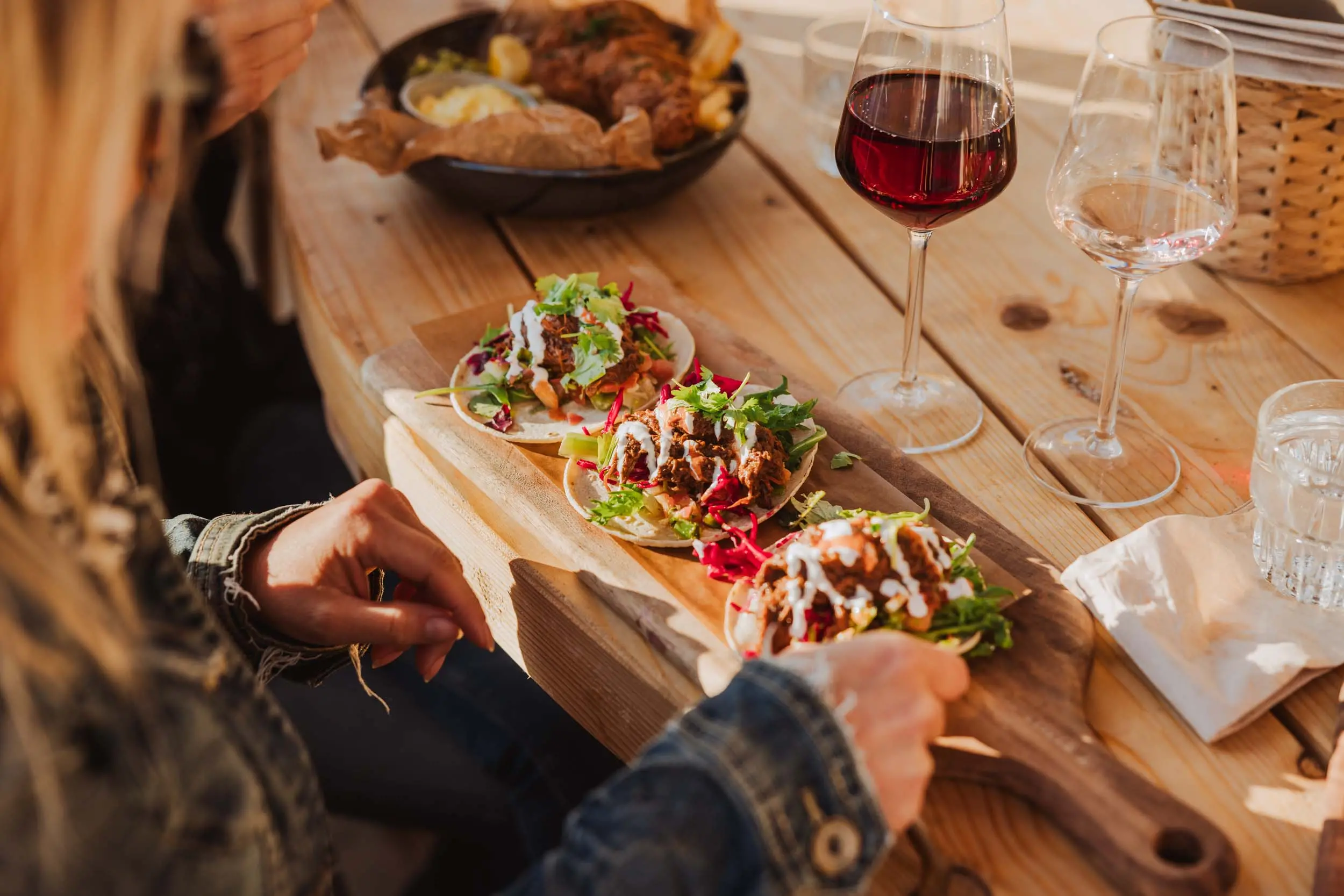 Tre stycken mjuka street-tacos på en bräda på ett bord med en tjej som precis ska ta upp en hos restaurang Monique.