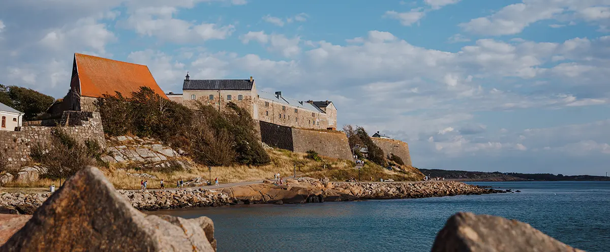 En fästningsbyggnad i sten som reser sig ovanför strandpromenad och hav, sedd från havet.